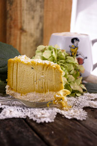 A slice of yellow coloured Coconut cheesecake with a fork slicing into it on a white cloth with green hydrange flowers in the background and a mug with an illustration of two children playing leap frog on the front against a wooden backdrop