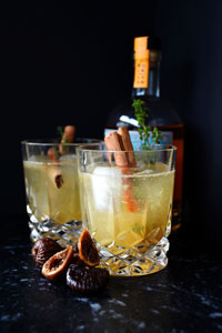 Two yellow coloured cocktails with cinnamon sticks and rosmary garnish showing out of the top in a short crystal glass against a black backdrop with dried figs sitting in front and a liquor bottle in the background