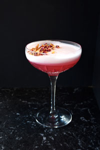 A pink cocktail with white foam and dried flower petals on top in a crystal cocktail glass against a black background