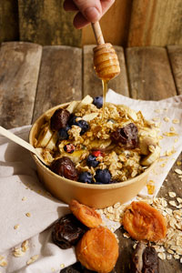 A bowl of overnight oats with mixed fresh and dried fruits sitting on a neutral coloured napkin on a wooden table and a hand holding a wooden honey dipper drizzling honey over the bowl