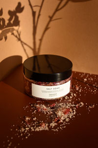 A container of bath salts with some of the salts spread across the foreground and a shadow of flowers in the background